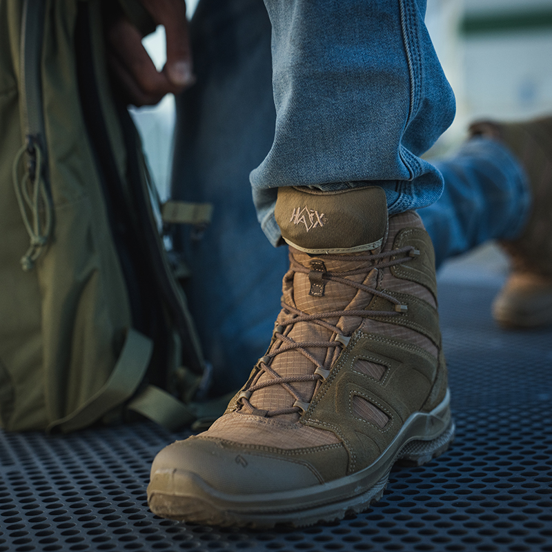 Close up of Black Eagle Athletic Mid Coyote worn with jeans as a person kneels beside an olive drab backpack