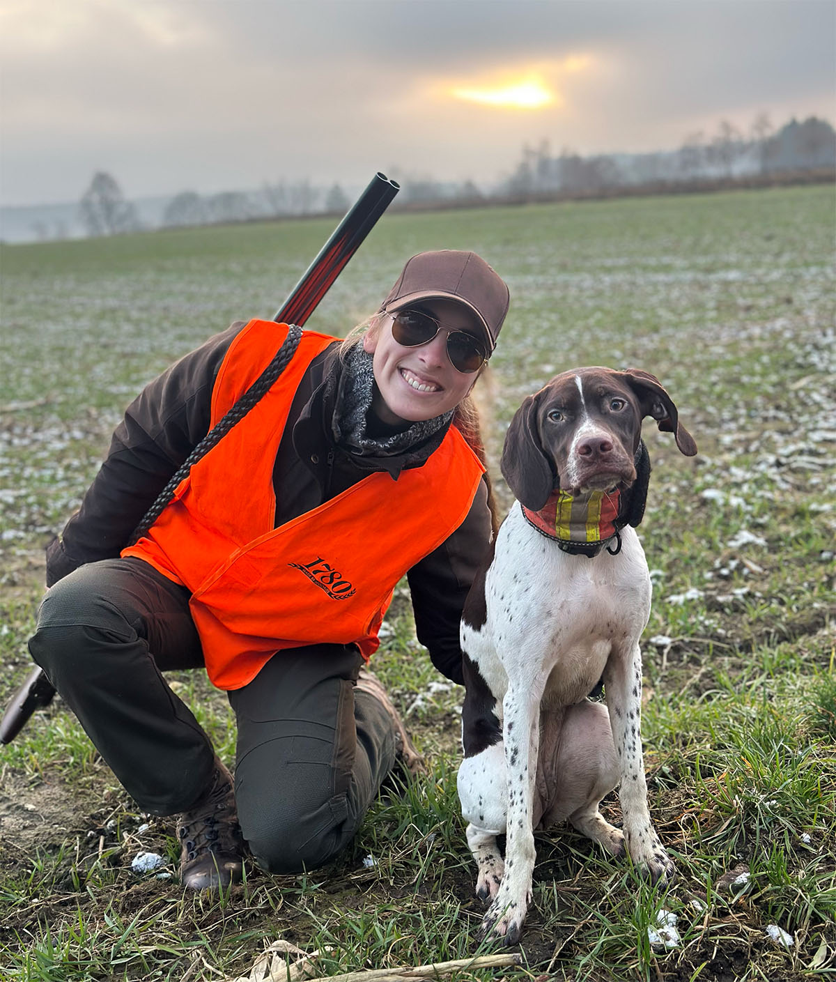 A female hunter in HAIX boots and orange vest poses with her pointer dog in a field during a sunset hunting trip.