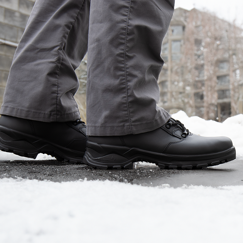 Close up of black Enforce X High Winter boots worn with grey pants, standing on snow-covered blacktop