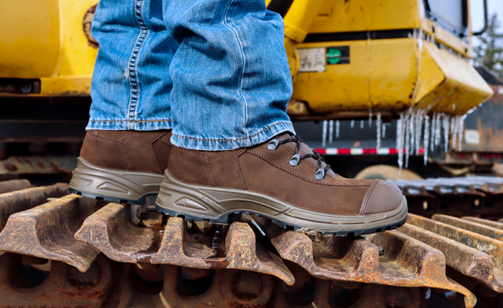 A person wearing durable brown HAIX work boots standing on the metal tracks of a yellow excavator in winter weather.